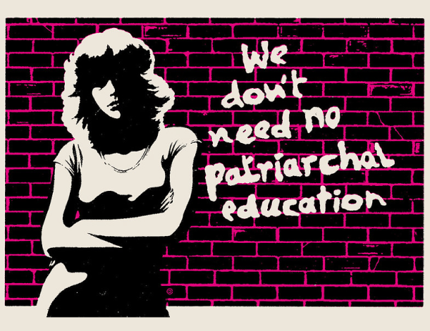 Illustration of a woman with folded arms, right in front of a brick wall. On the wall, graffiti that says We don't need no patriarchal education
