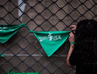 A person with long, wavy hair ties a green bandana to a wire fence. The bandana says "Aborto Legal", which means Legal Abortion. Green is the color for safe and legal abortion movements in Latin America.