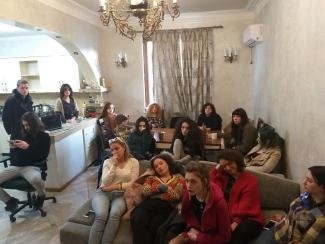 This photo represents a group of 15 workers sitting together in a living room with white walls. Some are sleeping, others are standing, talking with each other, or watching their phones.