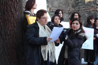 The photo on depicts eight women standing together during a protest. Many are holding banners while Sopo is holding the megaphone close to the mouth of a woman worker with short red hair, wearing a white scarf and a black coat reading a manifesto.