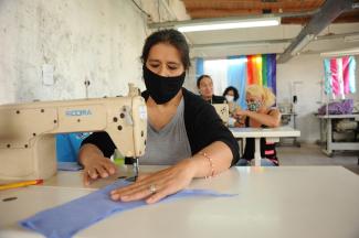 Depicts trans and travesti women behind sewing machines, with a close-up of a person with dark hair, wearing a black surgical mask, sowing a baby blue piece of fabric. The curtains in the workshops are the color of the LGTBQIA+ and trans flags.