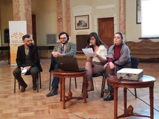 This photo represents Sopo speaking at a public event indoors. She is holding the microphone while reading her notes and sitting on a chair between three other people who are either speakers or moderators. 