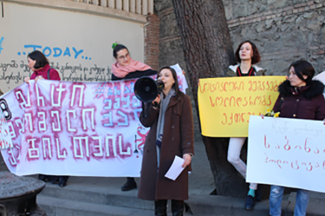 This photo shows four people with posters at a protest and in the middle a woman with a megaphone speaking