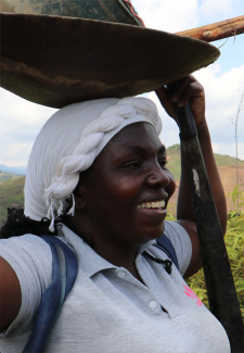 Black woman in a white headscarf and gray t-shirt, she is smiling and is holding a vessel above her head