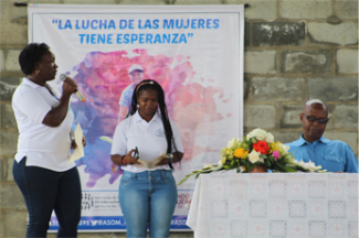 Black woman speaking in a mic, there is a black girl standing next to her and a man sitting at the table