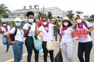 Photo of five black women in the front on a demonstration, wearing facemasks and holding posters
