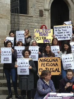 Photo shows shows four people with posters at a protest and in the middle a woman with a megaphone speaking. 