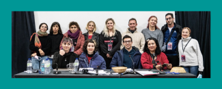 Un panel de 13 personas de pie detrás de una silla de conferencia. Sobre la mesa hay hojas de papeles, micrófonos y botellas de agua. Detrás de ellos se puede ver una pared blanca y cortinas negras.