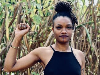 Picture of an African woman in a field, wearing her locs in a bun. She is holding her right fist up, and on each knuckle as well as her chin there are stripes with the colors of the pride flag.
