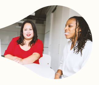 The image shows the image of two women sitting in a work setting. The one to the left has a red blouse and shoulder length brown hair. The one to the right is wearing a white blouse and has shoulder length micro dreadlocks.