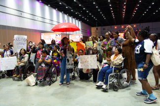 The photo shows a moment at the 15th AWID International Forum. There are disabled feminists (using mobility aids, like wheelchairs), deaf activist and sex work activists holding a red umbrella. 