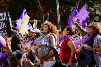Vibrant street march showcasing unity and feminism under purple flags. The woman in the forefront has a baby in a carrier, close to her chest.